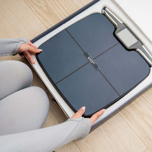 Person opening a box containing a digital scale with a person's hand visible.