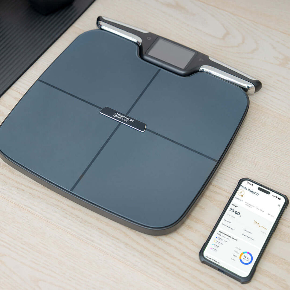 Smart weight scale on a wooden surface with a smartphone displaying app.