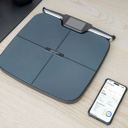 Smart weight scale on a wooden surface with a smartphone displaying app.
